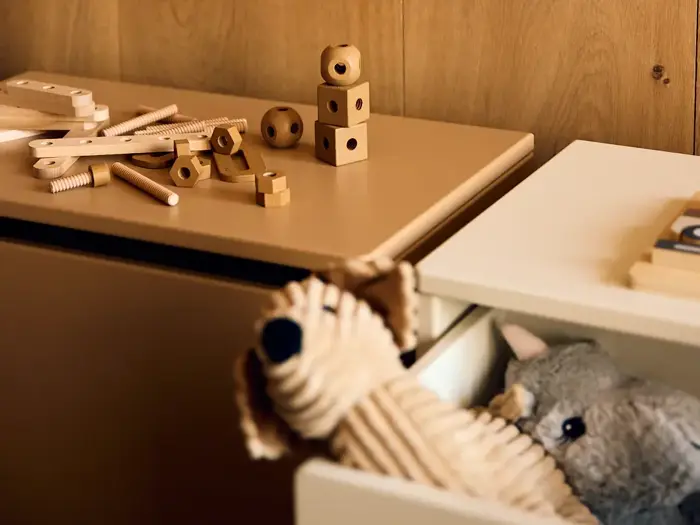 Legetøjsbolte og -klodser af træ ligger på et brunt bord. I en skuffe nedenunder ligger en udstoppet hund og et gråt plysdyr på en trævæg som baggrund.