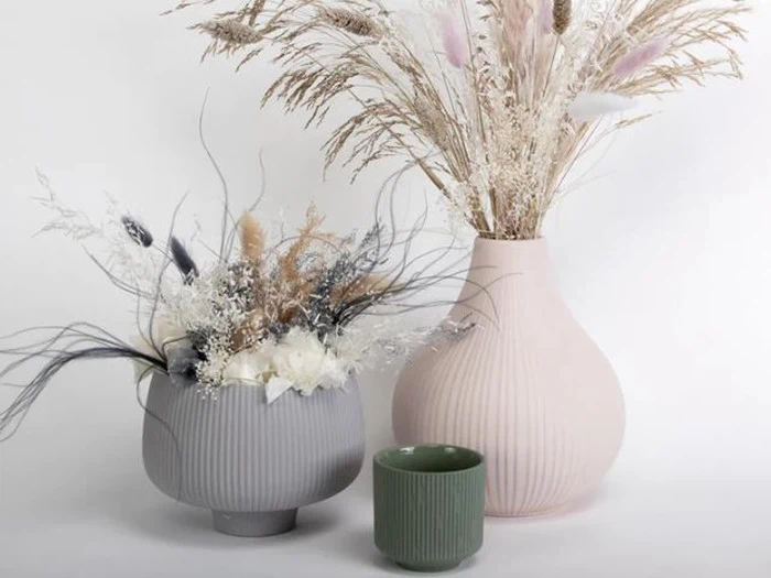 Tre vaser med tørrede blomster: en grå vase og en pink vase indeholder forskellige tørrede græsser; en lille grøn vase er tom foran dem, alt sammen på en almindelig lys baggrund.