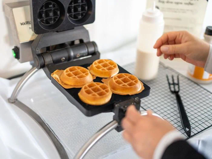En person laver fire runde vafler i et vaffeljern, mens en flaske med dej og redskaber står synligt på en rist i nærheden.