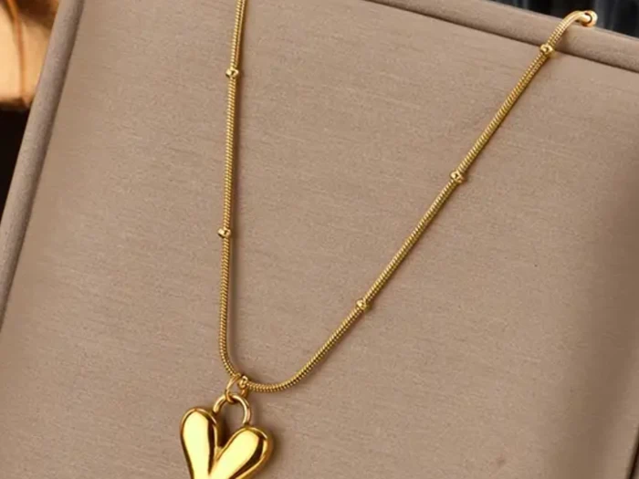En guldhalskæde med en fin kæde og et skinnende, stiliseret hjerteformet vedhæng på et beige smykkestativ.