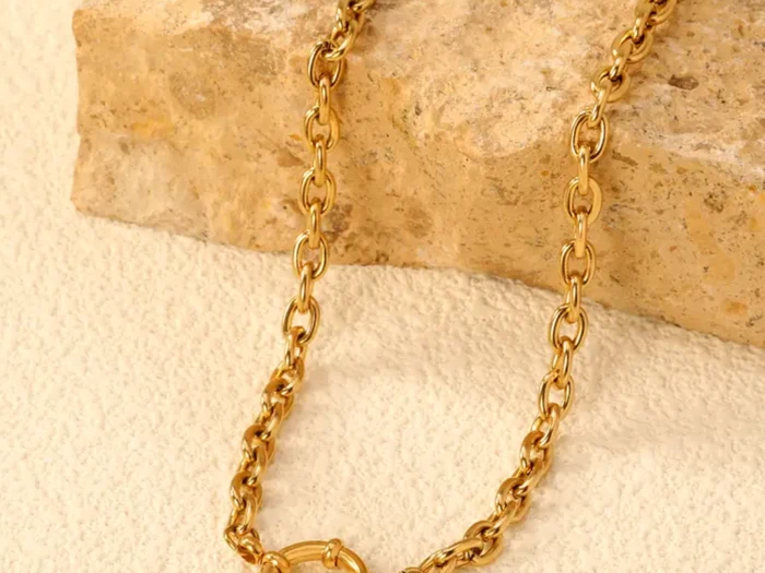 En guldkæde med ovale led vises på en struktureret beige overflade, draperet over en rå sten.