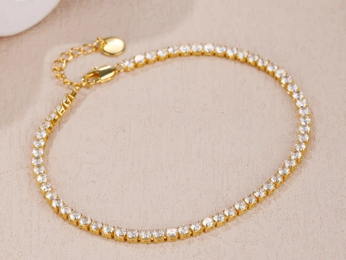 Et delikat guldarmbånd med klare runde ædelsten jævnt fordelt langs båndet, vist på en beige tekstureret overflade.