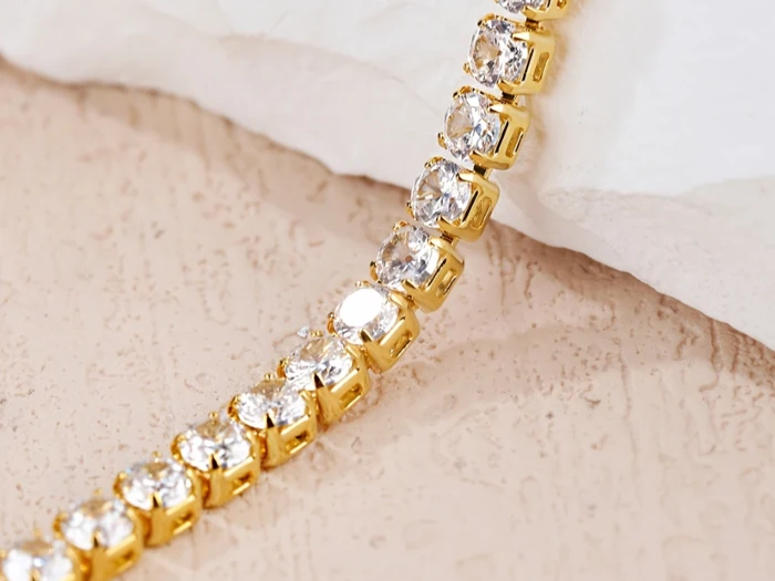 Nærbillede af et guldarmbånd med runde, klare ædelsten indfattet i enkelte led, vist på en struktureret beige overflade.