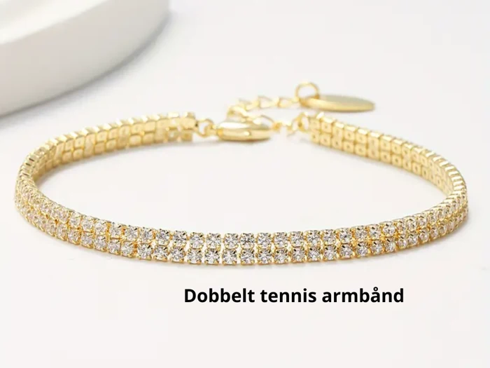 Et dobbeltradet tennisarmbånd i guld med funklende sten og en justerbar lås, vist på en lys overflade. Teksten lyder: Dobbelt tennis armbånd.