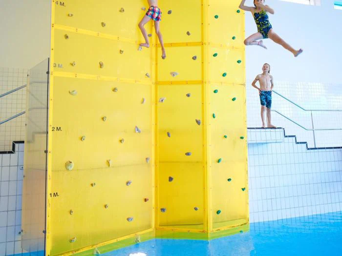 Tre børn i badetøj er i en indendørs pool. To klatrer eller springer fra en gul klatrevæg over vandet, mens en anden står på en trappe ved siden af poolen og ser på.