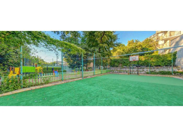 En udendørs sportsbane med grønt gulv, en basketballkurv, hegn, en legeplads og træer i baggrunden på en solrig dag.