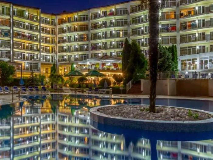 Et stærkt oplyst hotel i flere etager om natten afspejles i en rolig swimmingpool med synlige palmer, grønne områder og udendørs siddepladser.