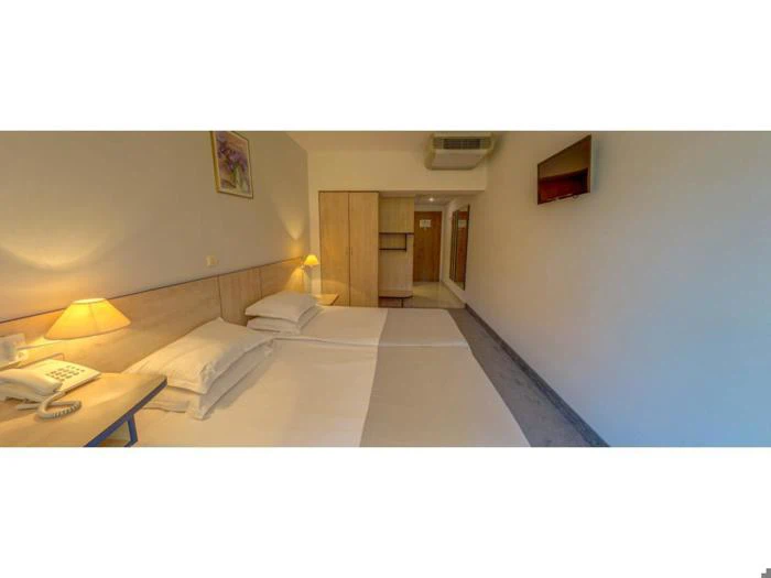 Et hotelværelse med to enkeltsenge, et natbord med lampe og telefon, et træklædeskab, væghængt tv og et maleri over hovedgærdet.