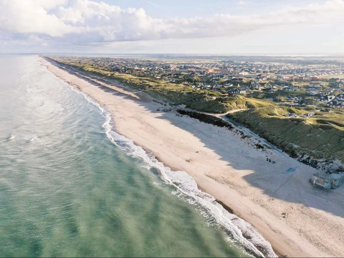 Luftfoto af en lang sandstrand omgivet af blide bølger på den ene side og græsklædte klitter med huse på den anden, under en delvis overskyet himmel.