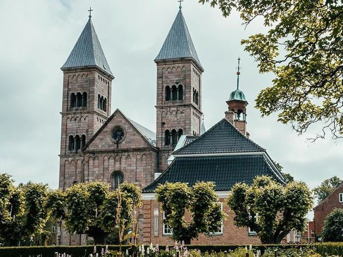 En historisk stenkirke med to høje tårne, buede vinduer og et mørkt tegltag, omgivet af grønne træer og en overskyet himmel.