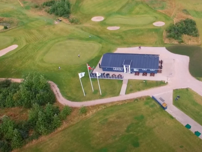 Luftfoto af en golfbane med et klubhus, flere flag, grønne fairways, sandbunkere og nogle få golfspillere på banen.