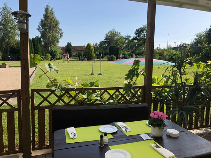 Et dækket spisebord på en overdækket terrasse har udsigt til en grøn græsplæne, legeplads og have på en solrig dag.