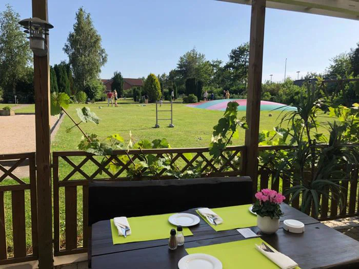 Et dækket spisebord på en overdækket terrasse har udsigt til en grøn græsplæne, legeplads og have på en solrig dag.