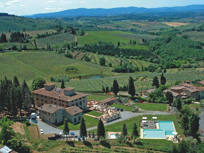 Luftfoto af en toscansk villa med pools, haver og omkringliggende bølgende grønne bakker, vinmarker og cypresser under en blå himmel.