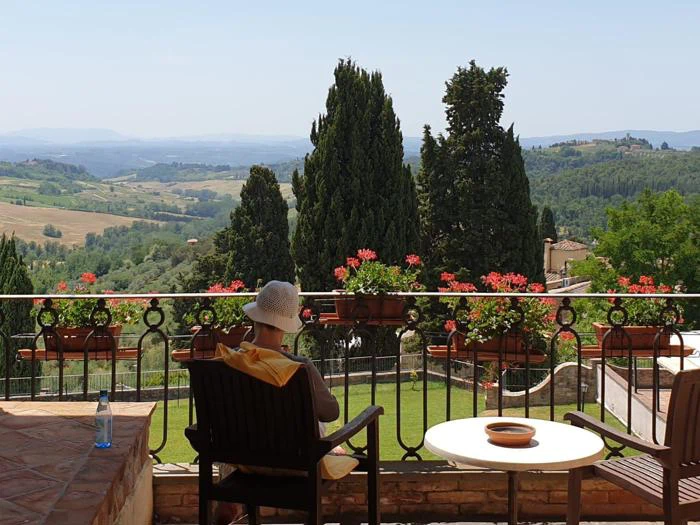 En person med solhat sidder på en terrasse med blomster og udsigt over et naturskønt landskab med bølgende bakker og træer under en klar himmel.
