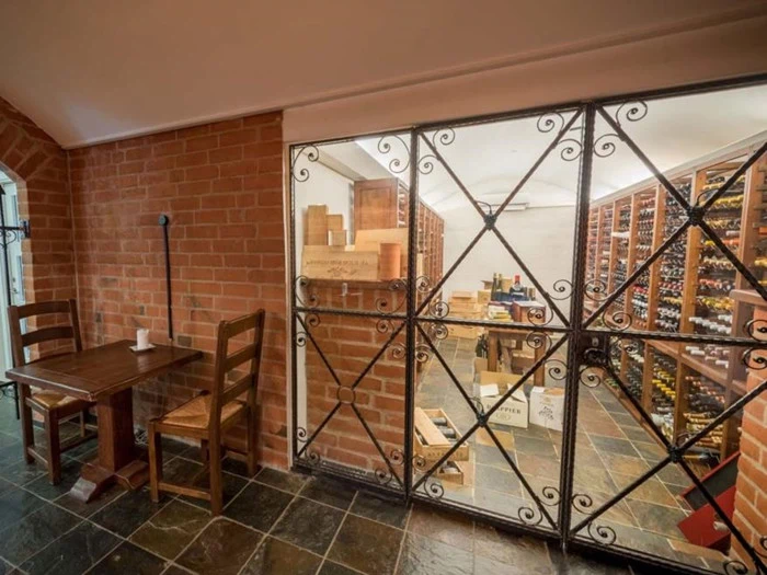 En vinkælder med murstensvægge, hylder fyldt med vinflasker bag en smedejernslåge og et træbord med to stole i forgrunden.