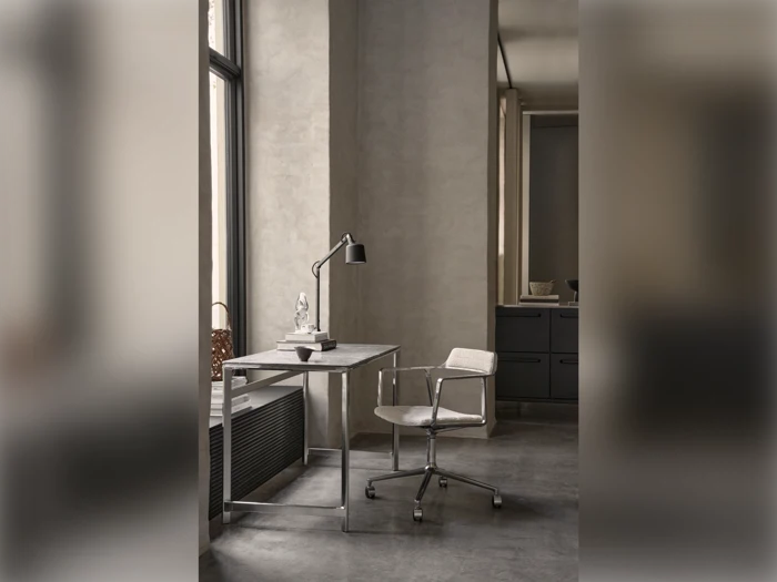 Minimalistisk arbejdsplads med metalskrivebord, justerbar lampe, stol på hjul og pyntegenstande ved et stort vindue i et neutralt rum.