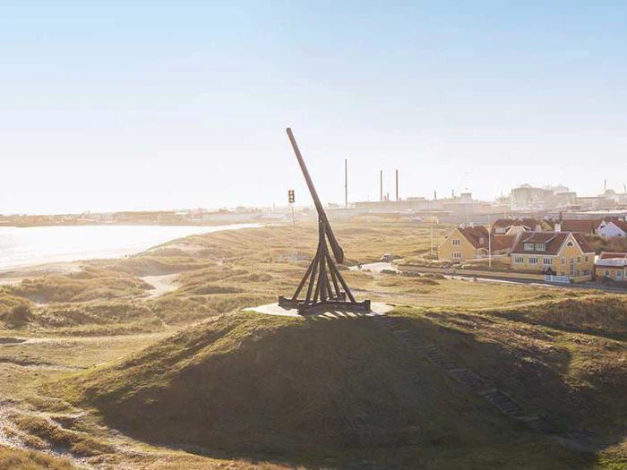 En stor trebuchet af træ står på en græsklædt bakke nær kysten med huse og et industriområde i baggrunden under en klar himmel.