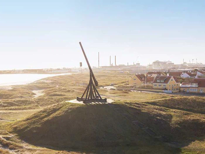 En stor trebuchet af træ står på en græsklædt bakke med udsigt over en kystby med huse og industribygninger i baggrunden under en klar himmel.