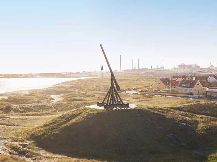 En stor trebuchet af træ står på en græsklædt bakke med udsigt over en kystby med farverige huse og et industriområde i baggrunden.