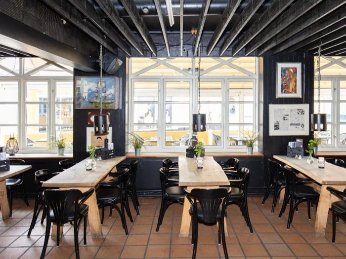 Moderne restaurantinteriør med træborde, sorte stole, hængelamper og store vinduer, der lukker naturligt lys ind; planter og kunstværker pynter i lokalet.