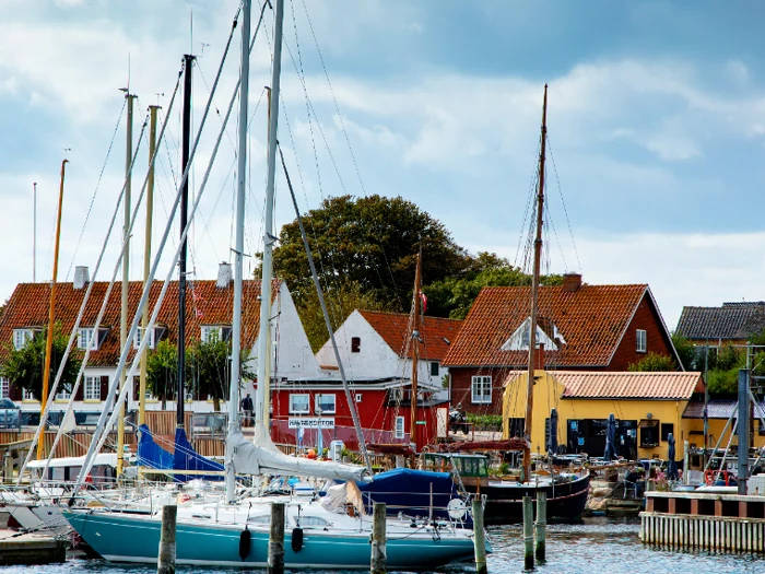 Farverige sejlbåde lægger til ved en marina med charmerende røde og gule huse og frodige træer i baggrunden under en delvis overskyet himmel.