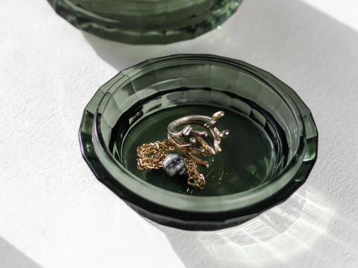 Et grønt glasfad med en guldkæde med en sort og hvid perle og en grenformet sølvring, placeret på en hvid overflade.