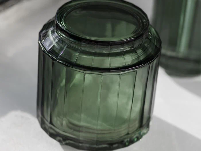 Nærbillede af en tom, mørkegrøn glaskrukke, der ligger på hovedet på en hvid flade og viser dens strukturerede, lodrette riller og flade bund.