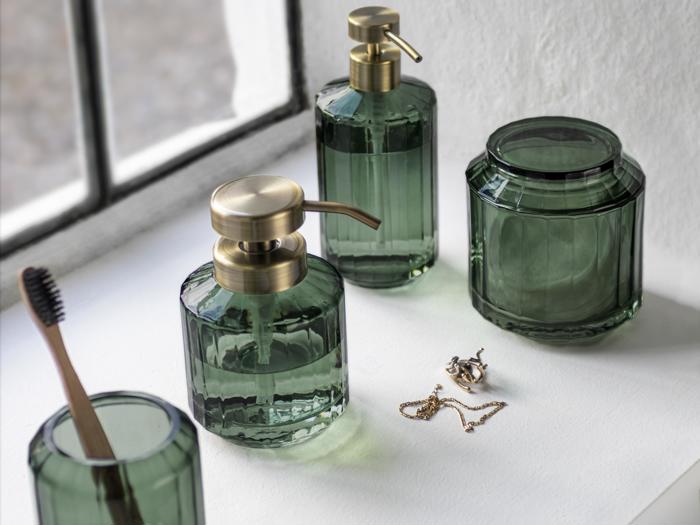 Badeværelsestilbehør i grønt glas med guldfarvede pumper og låg er anbragt i en hvid vindueskarm sammen med en tandbørste, en halskæde og et par øreringe.