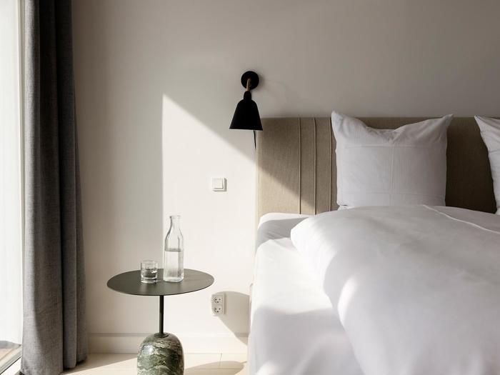 Et moderne, minimalistisk soveværelse med en pænt redt seng, et rundt sidebord med et glas og en vandflaske og en sort væglampe over, oplyst af naturligt sollys.