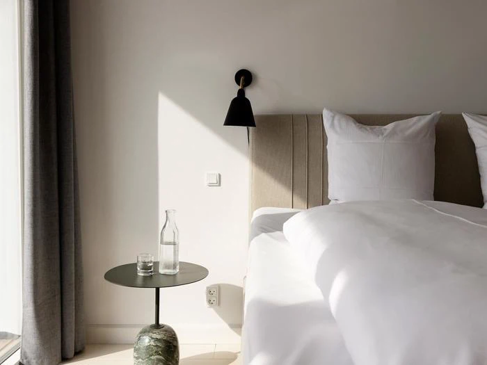 Et moderne, minimalistisk soveværelse med en pænt redt seng, et rundt sidebord med et glas og en vandflaske og en sort væglampe over, oplyst af naturligt sollys.