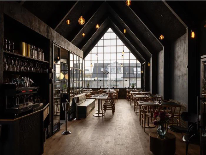 Moderne, svagt oplyst café med træborde og -stole, højt hvælvet loft, store gittervinduer og hængende pendellamper, som skaber en hyggelig og stilfuld atmosfære.