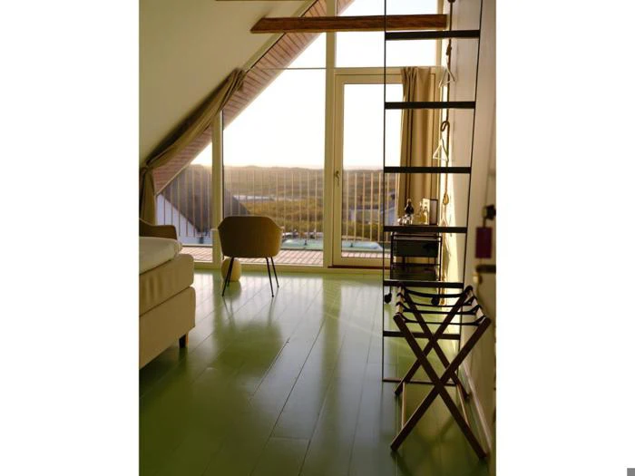 Et lyst, moderne værelse med grønne gulve, en seng, en stol og et stort vindue med udsigt til en naturskøn udendørs udsigt ved solnedgang.