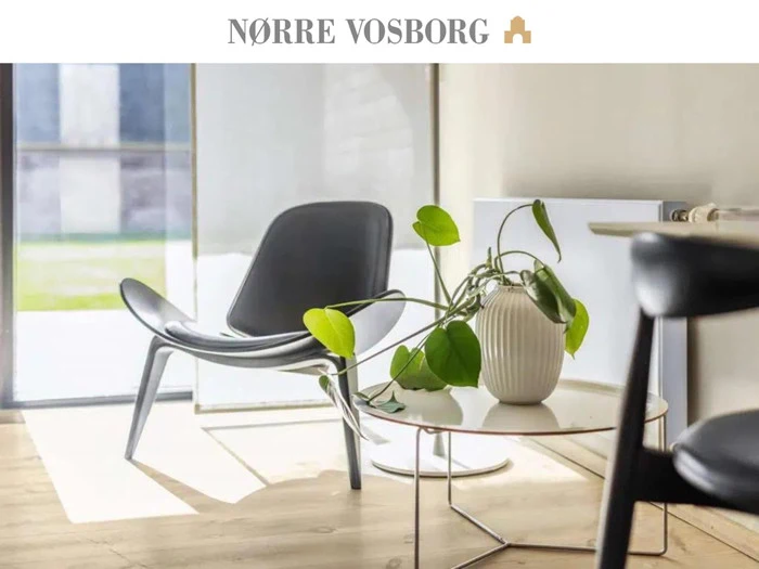 En moderne loungestol og et rundt bord med en potteplante i et solbeskinnet, minimalistisk rum. Teksten "NØRRE VOSBORG" står øverst.