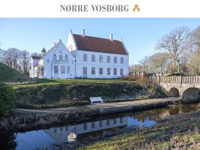 En stor hvid historisk herregård, Nørre Vosborg, ligger ved en voldgrav med en stenbro, omgivet af træer og et græsklædt landskab under en klar blå himmel.