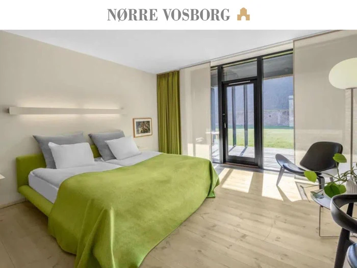 Moderne hotelværelse med en dobbeltseng dækket af grønt sengetøj, store vinduer med gennemsigtige gardiner, en stol og lyst trægulv. Sollyset strømmer ind i rummet.