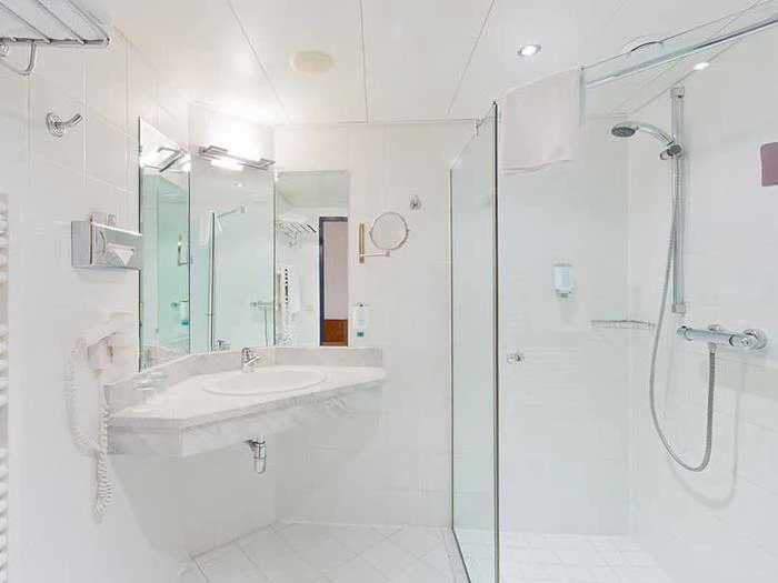 Moderne hvidt badeværelse med bruseafskærmning i glas, vægmonteret brusehoved, stort spejl, håndvask, hårtørrer og håndklædetørrer, alt sammen med god belysning og rene overflader.