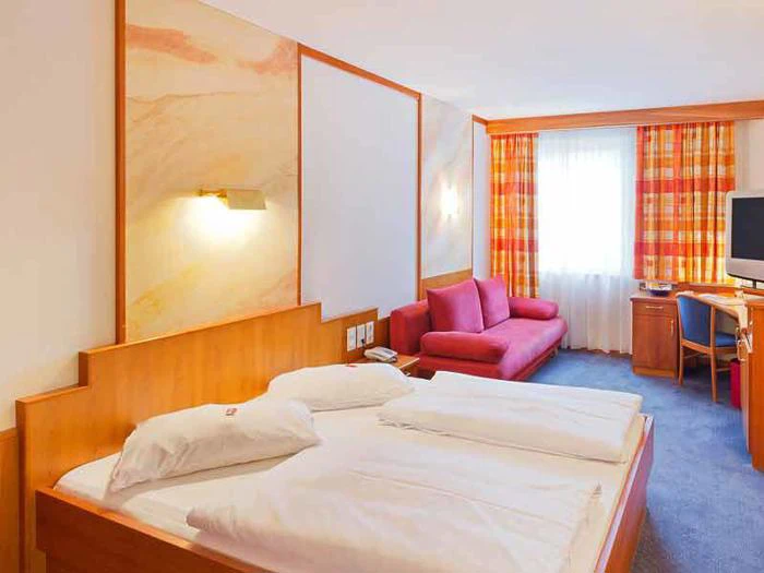 Et hotelværelse med dobbeltseng, rød sofa, skrivebord, tv og et vindue med orange ternede gardiner. Væggene og møblerne er af lyst træ, og værelset er stærkt oplyst.