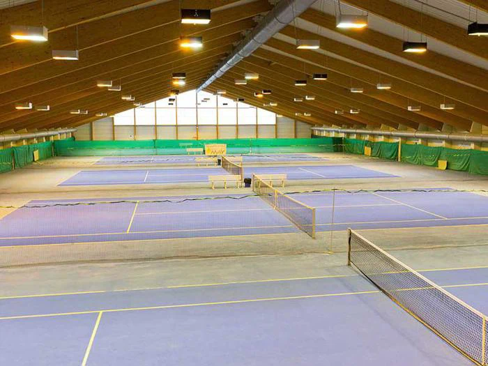 Stort indendørs tennisanlæg med flere blå baner, grønne vægge og et højt træloft, oplyst af ovenlys.