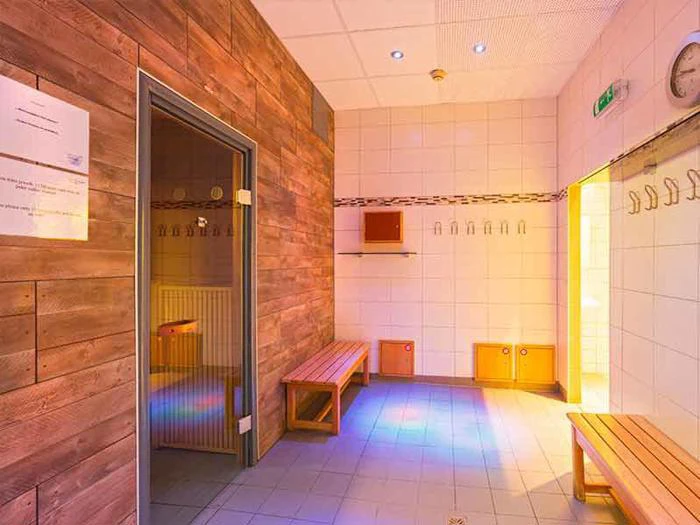 Et lyst, flisebelagt omklædningsrum med træbænke, vægkroge, et ur og en åben dør, der fører til en sauna beklædt med træpaneler.