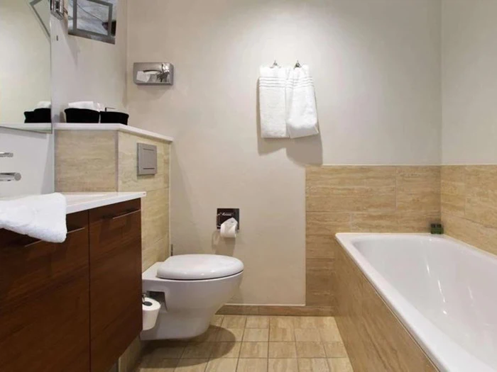 Moderne badeværelse med væghængt toilet, hvidt badekar, træskabe, klinkegulv og -vægge samt to hvide håndklæder på væggen.