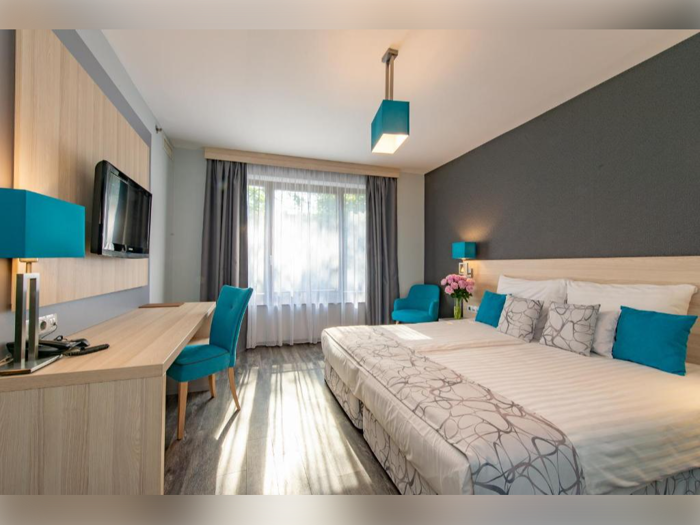 Moderne hotelværelse med en stor seng, blågrønne detaljer, skrivebord, stol, lænestol, fladskærms-tv og et vindue med gennemsigtige gardiner, der lukker naturligt lys ind.