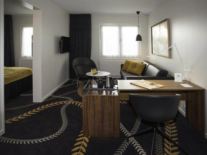 Moderne hotelværelse med mørkt mønstret gulvtæppe, skrivebord og stol i træ, sofa i gule nuancer, lænestol og stort vindue med gardiner, der lukker naturligt lys ind.
