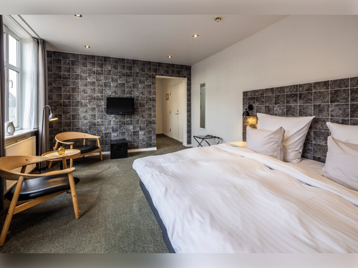 Moderne hotelværelse med en stor seng, to træstole, et lille bord, væghængt tv og struktureret mørk accentvæg.