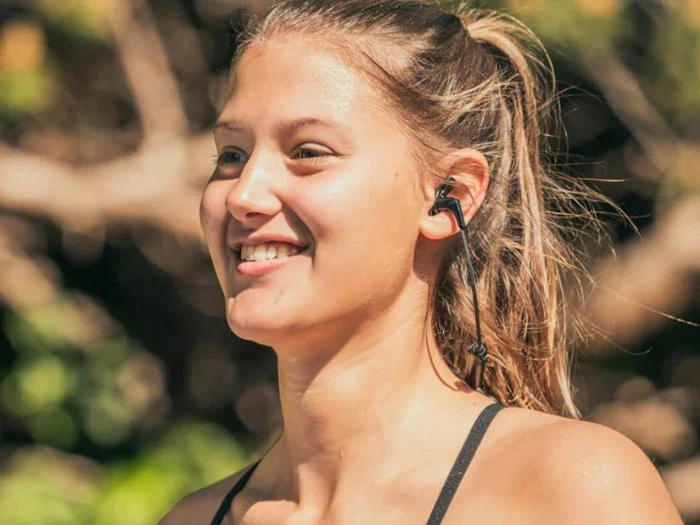 En ung kvinde med hestehale smiler, mens hun har trådløse øretelefoner og sportstøj på udendørs i solskinsvejr.