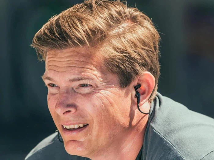 En mand med lysebrunt hår, iført høretelefoner, sveder og ser ud til at anstrenge sig, muligvis under træning, i stærkt sollys.
