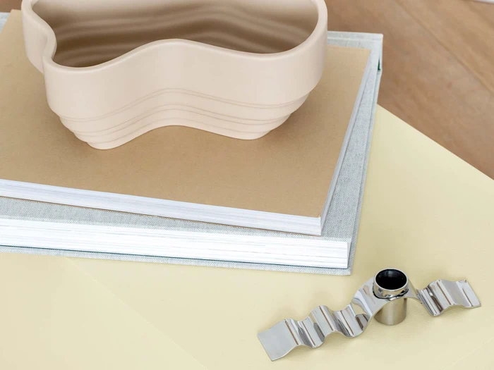 En bølget beige skål står oven på to stablede bøger ved siden af et bølget sølvtilbehør på en lysegul overflade.