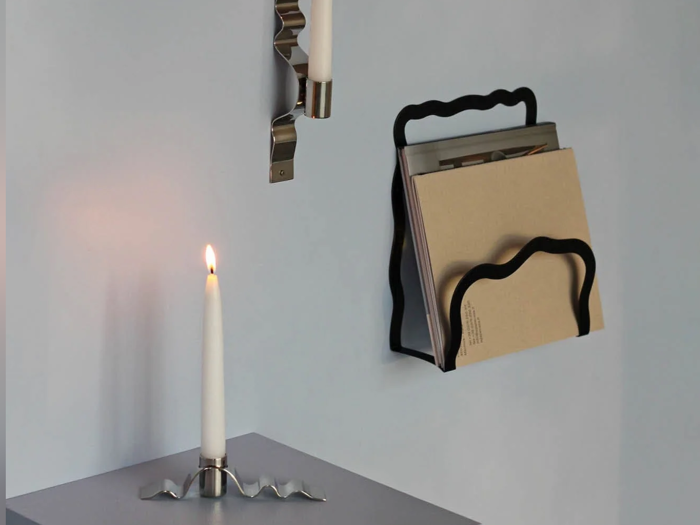 Et brændende hvidt stearinlys i en bølget metalholder står på et bord; over det er der en vægmonteret sort bølget reol med bøger eller magasiner ved siden af en anden lysestage.