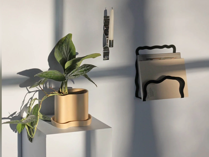 En potteplante på en piedestal ved siden af en væghængt magasinholder med mapper og en penneholder, alt sammen i blødt sollys.