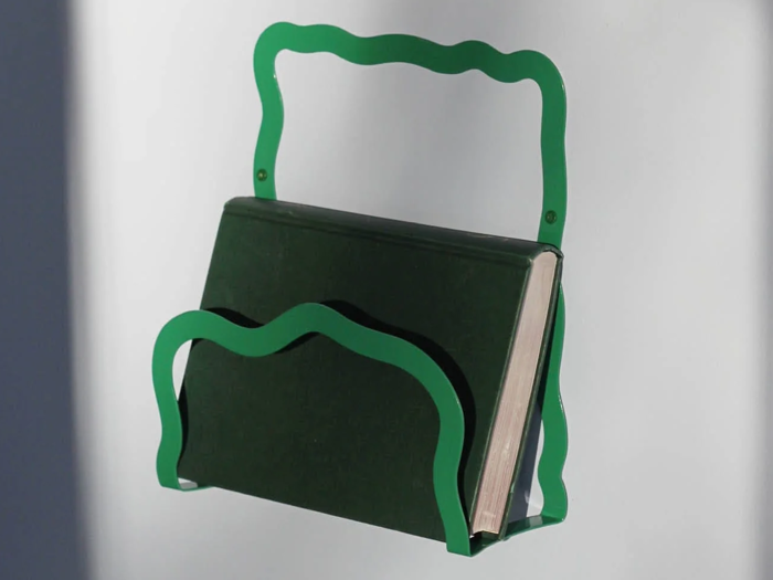 En grøn, bølgekantet væghængt metalreol holder en mørkegrøn bog oprejst mod en hvid baggrund.